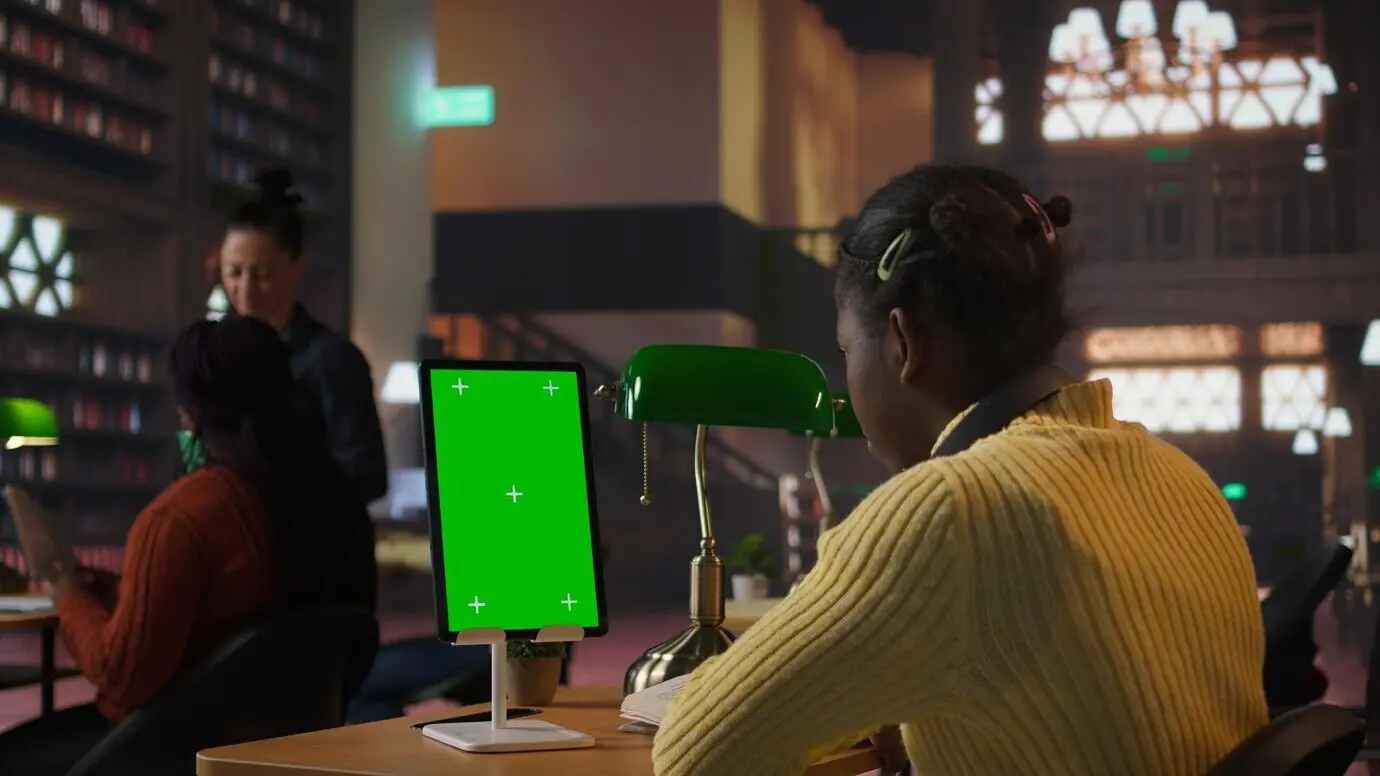 Eine afroamerikanische Schülerin schreibt in einer Bibliothek neben einem Chroma-Key Unterrichtsnotizen.